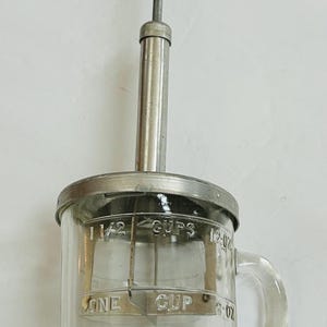 May include: A vintage glass measuring jug with a metal lid and handle. The jug has measurements for 1/4, 1/2, 3/4, and 1 cup, and 59, 118, 177, and 237 ml. A metal plunger with a red knob is inserted into the jug.