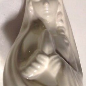 Op de afbeelding: Witte keramische figuur van een vrouw met haar handen gevouwen in gebed. De vrouw draagt een lange jurk en heeft een serene uitdrukking op haar gezicht.
