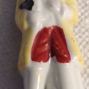 May include: A porcelain figurine of a person wearing a yellow coat, red pants, and a white shirt. The figurine is standing on a white base with a gold line.