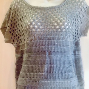 May include: A gray, short-sleeved sweater with a round neckline. The top portion features an open, lacy knit design, while the lower section has a horizontal ribbed pattern. The sweater appears to be made of a soft, possibly shimmering material.