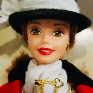 May include: A doll with a fair complexion, wearing a black and white hat with a white bow. She has red lips, gold earrings, and a white scarf with a gold key-shaped pendant. The doll is dressed in a red and black outfit.