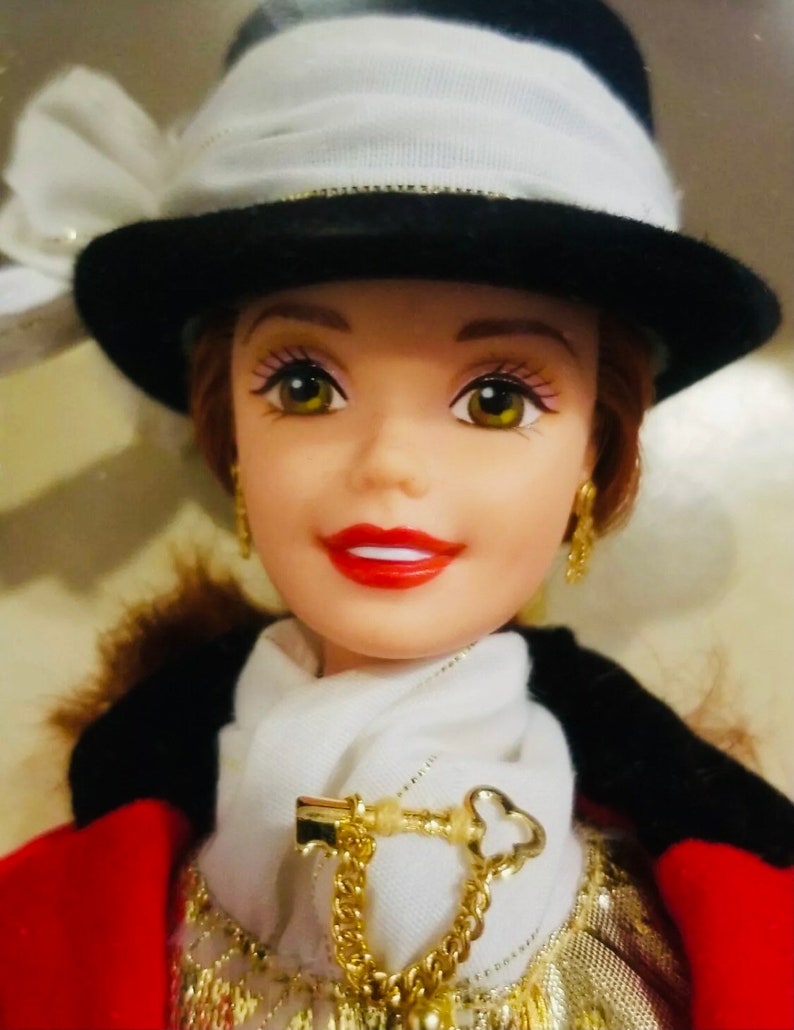 May include: A doll with a fair complexion, wearing a black and white hat with a white bow. She has red lips, gold earrings, and a white scarf with a gold key-shaped pendant. The doll is dressed in a red and black outfit.