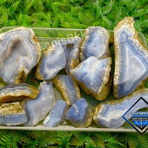 May include: A collection of blue agate stones with brown banding in a clear plastic container. The stones are various shapes and sizes. The container has the text "LAPISBK LAPIS LAZULI CO. LTD" on the bottom.