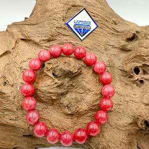 Op de afbeelding: Een armband met grote, ronde, roodroze edelstenen. De armband wordt gepresenteerd op een stuk getextureerd, lichtbruin hout. Een blauw-wit logo is zichtbaar op de achtergrond.