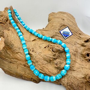 May include: A turquoise beaded necklace with white detailing. The necklace features barrel-shaped beads strung together. The beads are a bright blue colour with white marbling. The necklace is displayed on a piece of driftwood.