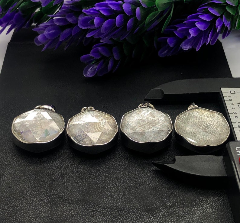 May include: Four silver pendants with a white, iridescent, and metallic finish. The pendants are round and have a geometric design. They are arranged in a row on a black surface.
