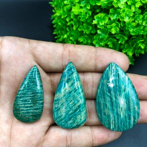 May include: Three teardrop-shaped green amazonite cabochons with a striated pattern. The stones are arranged in a row on a hand.