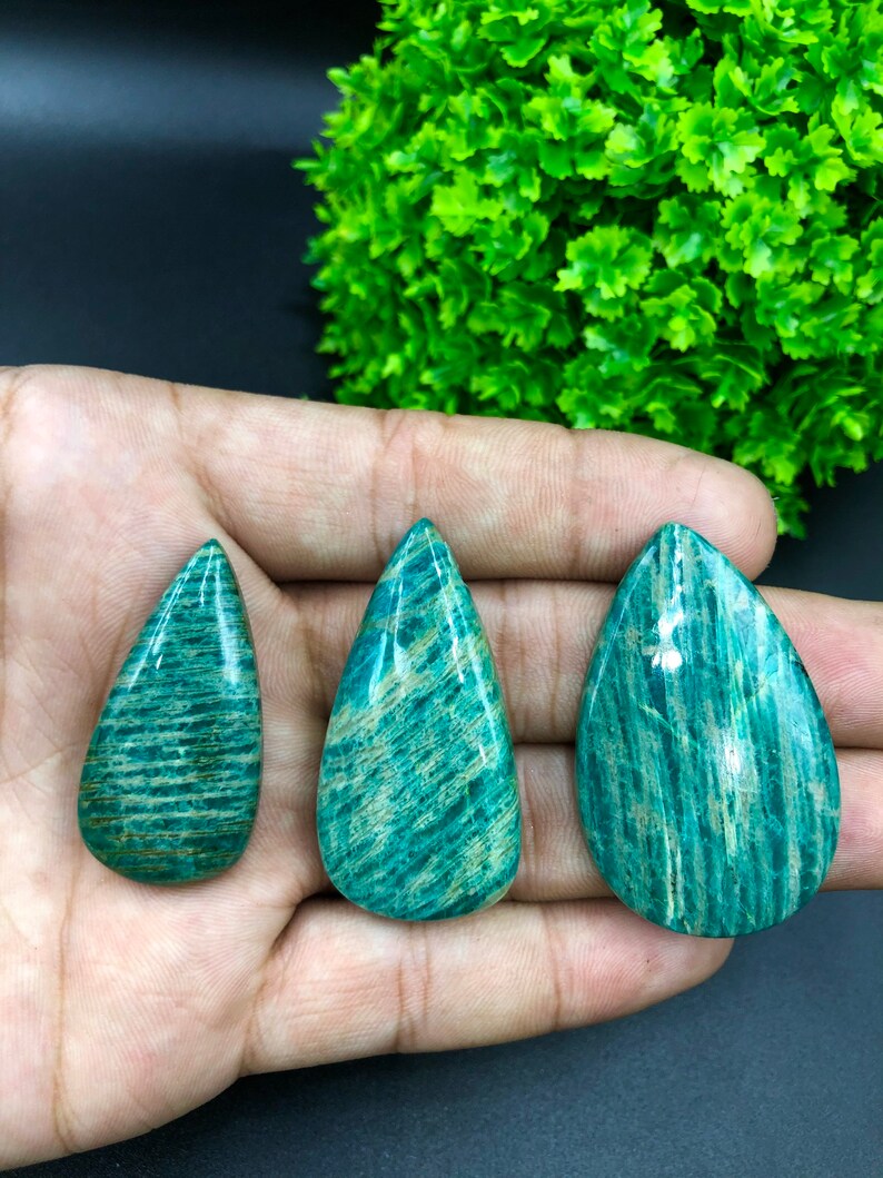 May include: Three teardrop-shaped green amazonite cabochons with a striated pattern. The stones are arranged in a row on a hand.