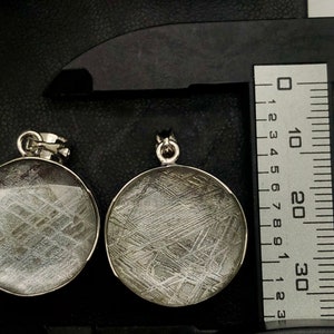 May include: Two silver pendants with a circular, clear glass front, showcasing a pattern of silvery-grey lines resembling a meteorite. The pendants are measured with a ruler and a digital scale.