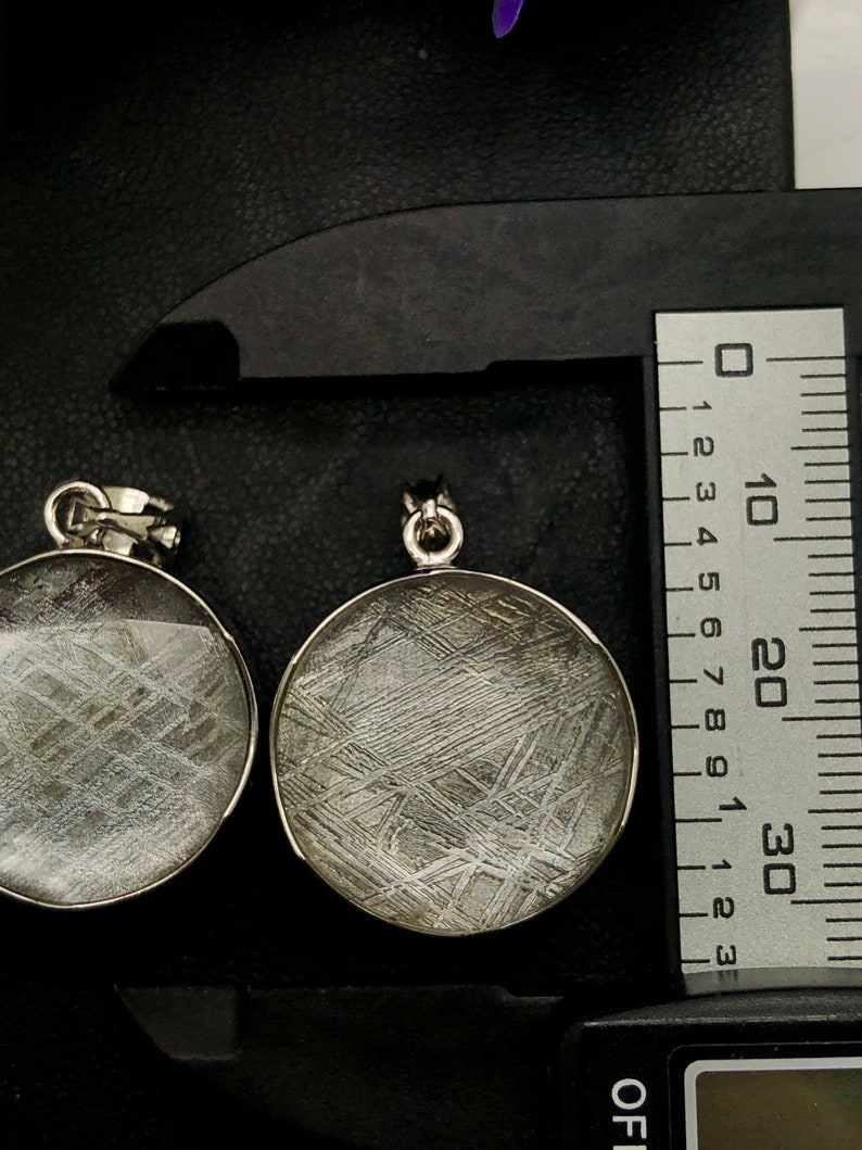May include: Two silver pendants with a circular, clear glass front, showcasing a pattern of silvery-grey lines resembling a meteorite. The pendants are measured with a ruler and a digital scale.