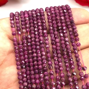 May include: A strand of ruby beads, faceted and strung on a pink thread. The beads are a deep, rich red color and have a slightly irregular shape.