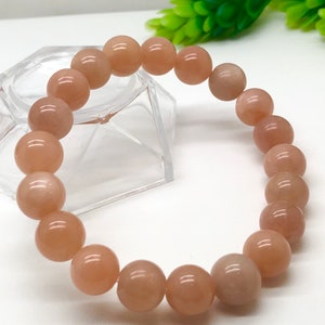 Puede incluir: Un brazalete hecho de cuentas de piedra de sol de color melocotón. Las cuentas son redondas y lisas, y el brazalete está encordado en un cordón elástico transparente.
