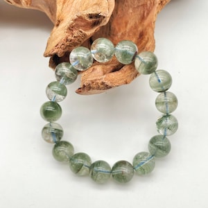 Könnte beinhalten: Ein Armband mit durchscheinenden grünen und klaren Quarz-Kugeln. Jede Kugel hat einen Durchmesser von etwa 2 cm und ist mit einem hellblauen Gummiband verbunden. Das Armband wird vor einem weißen Hintergrund mit einem Holzstück präsentiert.