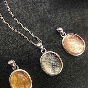 May include: Three silver pendants with a clear, oval-shaped glass containing a slice of meteorite. The meteorite has a unique, striated pattern. The pendants are on a black background.