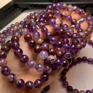 Könnte beinhalten: Eine Nahaufnahme eines mehrreihigen Armbands aus violetten und braunen Edelsteinperlen. Die Perlen sind rund und haben eine glatte, polierte Oberfläche. Das Armband besteht aus mehreren Perlenreihen, die miteinander verwoben sind, um ein einzigartiges und komplexes Design zu schaffen.