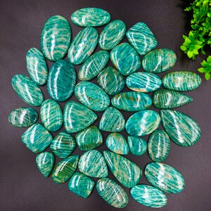 May include: A collection of 30 green amazonite cabochons with a striated pattern. The cabochons are various shapes and sizes, and are arranged in a semi-circle on a black background.