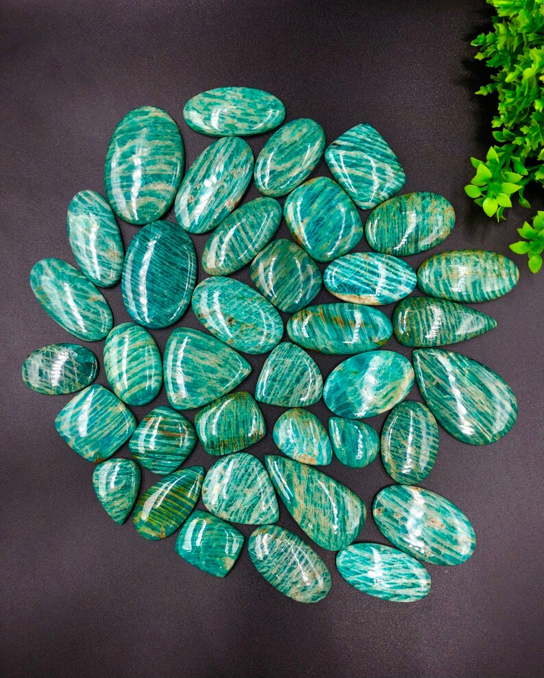 May include: A collection of 30 green amazonite cabochons with a striated pattern. The cabochons are various shapes and sizes, and are arranged in a semi-circle on a black background.