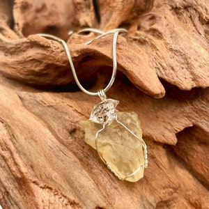 Puede incluir: Un colgante de cristal de citrino amarillo envuelto en alambre de plata, colgando de una cadena de plata. El colgante se muestra en un trozo de madera flotante.