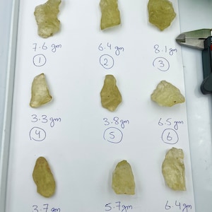Puede incluir: Nueve piezas de cristal amarillo-verde, cada una etiquetada con un número y un peso en gramos. Los cristales están dispuestos en tres filas de tres sobre una superficie blanca.