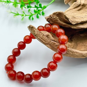 May include: A red-orange beaded bracelet with large, round beads. The bracelet is displayed on a piece of driftwood against a white background. The beads have a glossy finish and are strung together with a red cord.