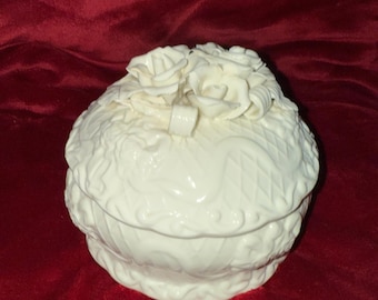 May include: A white ceramic trinket box with a lid, adorned with relief-style roses and intricate patterns. The box has a round shape and sits on a red surface. The overall design is elegant and decorative.