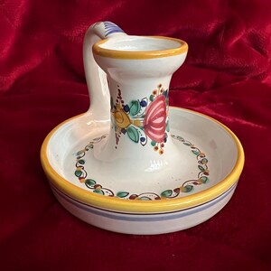 May include: White ceramic candle holder with a handle and a floral design in shades of red, yellow, green, and blue. The holder has a yellow rim and a blue and white stripe around the base.