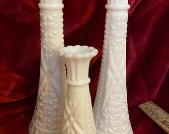 May include: Set of three white milk glass bud vases with a diamond pattern. The vases are different sizes and are arranged in a row on a red surface.