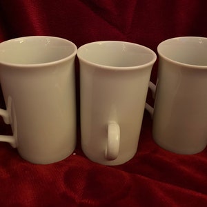 May include: Three white ceramic mugs with handles. The mugs are arranged in a row on a red surface.
