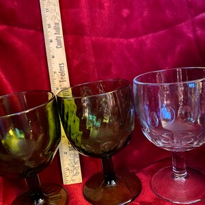 May include: Three stemmed drinking glasses, two are green glass and one is clear glass. The green glasses have a textured surface. The clear glass has a textured surface with small raised dots. A wooden ruler is in the background.