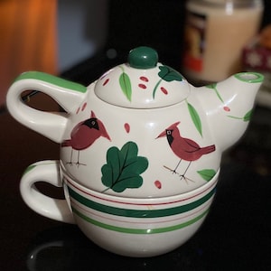 May include: White ceramic teapot and teacup set with a green, red, and white cardinal bird design. The teapot has a lid with a green knob. The teacup has a green and white striped band around the top.