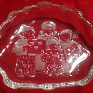 May include: A clear glass dish with a scalloped edge features an etched design of four carolers holding songbooks. The carolers are dressed in winter clothing and have hearts on their songbooks.