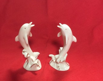 May include: Two white porcelain dolphin figurines with gold accents, leaping out of the water. The dolphins are facing each other and have a delicate, elegant design.