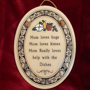 May include: A white oval ceramic plaque with a black floral border and a message that reads "Mom loves hugs Mom loves kisses Mom Really loves help with the Dishes". The plaque has a brown twine hanger and three small ceramic embellishments: a white flower, a green pear, and a red apple.