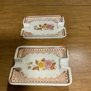 May include: Two white ceramic rectangular ashtrays with brown floral designs. The ashtrays have a raised rim and a small indentation in the center for cigarettes.