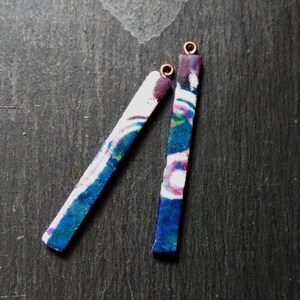 May include: Two rectangular earrings with a colorful, abstract pattern. The earrings are primarily blue, white, and purple, with a small gold-colored loop at the top. The earrings are displayed on a dark gray surface.