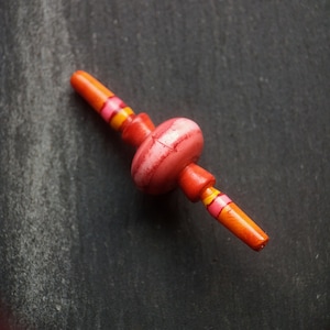 May include: A handcrafted, wooden bead with a central, rounded red section and orange ends. The orange sections are decorated with pink and yellow stripes. The bead is set against a dark, textured background.