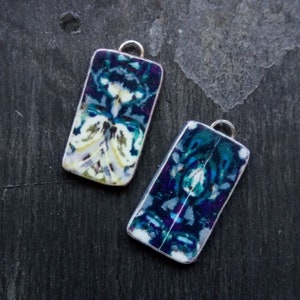 Puede incluir: Dos colgantes rectangulares de cerámica con un diseño abstracto azul, blanco y verde. Los colgantes tienen lazos plateados para colgar.