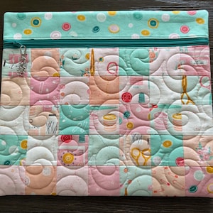May include: A rectangular zippered pouch featuring a patchwork design of pastel pink, mint green, and white fabric squares. The fabric is printed with sewing-themed motifs including scissors, buttons, and thread spools, finished with a teal zipper.