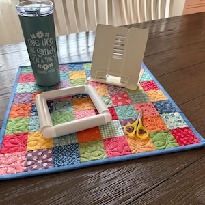 May include: A teal tumbler with the text "live life one stitch at a time" sits on a colorful patchwork quilt. A white embroidery hoop, a small pair of yellow scissors, and a white book stand are also on the quilt.