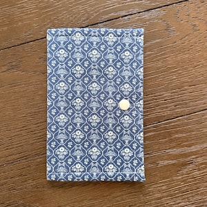 May include: A rectangular fabric case featuring a blue and white repeating pattern of floral baskets within a lattice design. The case is secured with a single white snap button centered on the right edge, with stitched borders along the perimeter.