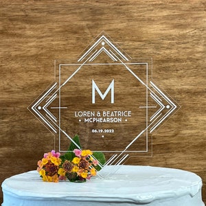 May include: Clear acrylic wedding cake topper with a geometric design. The topper features the letter "M" in the center, surrounded by a square frame with a geometric pattern. The text "Loren & Beatrice Mcphearson" and the date "06.19.2022" are also included.