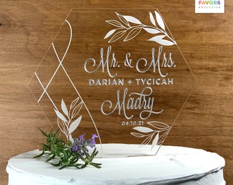 May include: A clear, geometric wedding cake topper with etched design and silver painted text. The topper reads "Mr. & Mrs. Darian + Tycicah Madry 04.10.21" with floral accents. The cake is white with a sprig of rosemary and lavender.