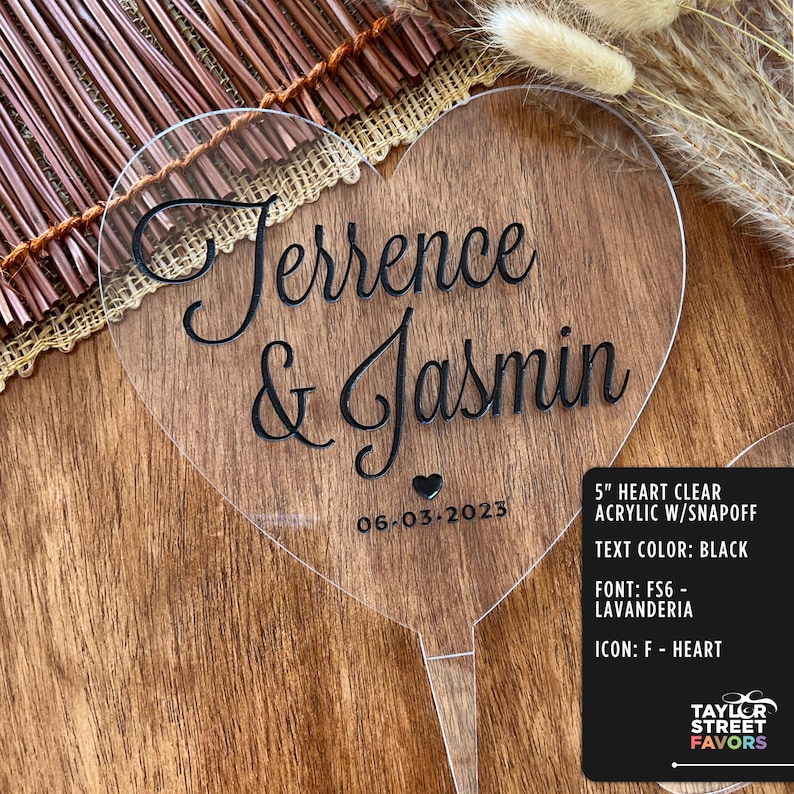 May include: A clear acrylic heart-shaped wedding cake topper with the names "Terrence & Jasmin" and the date "06-03-2023" in black script. The topper is 5 inches in size and has a snap-off design.