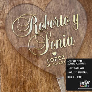 May include: A clear acrylic heart-shaped wedding favor with gold lettering. The names "Roberto y Sonia" are displayed, along with "LOPEZ" and the date "06.10.202". The text color is gold, and the font is FS11 Balmoral.