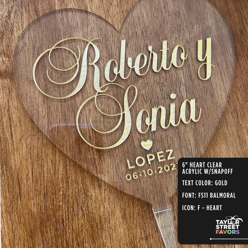 May include: A clear acrylic heart-shaped wedding favor with gold lettering. The names "Roberto y Sonia" are displayed, along with "LOPEZ" and the date "06.10.202". The text color is gold, and the font is FS11 Balmoral.