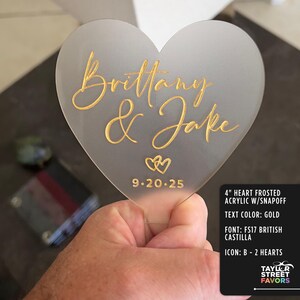 May include: A frosted acrylic heart-shaped wedding favor, 4 inches in size, with gold lettering that reads "Brittany & Jake" and the date 9.20.25. The design includes two heart icons. The text color is gold.