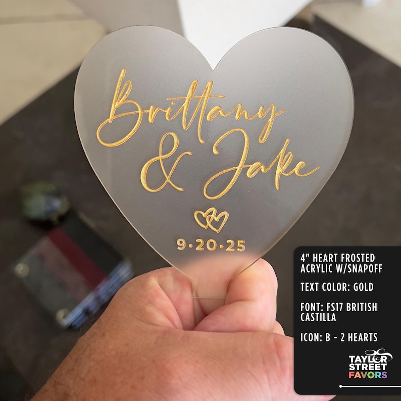 May include: A frosted acrylic heart-shaped wedding favor, 4 inches in size, with gold lettering that reads "Brittany & Jake" and the date 9.20.25. The design includes two heart icons. The text color is gold.