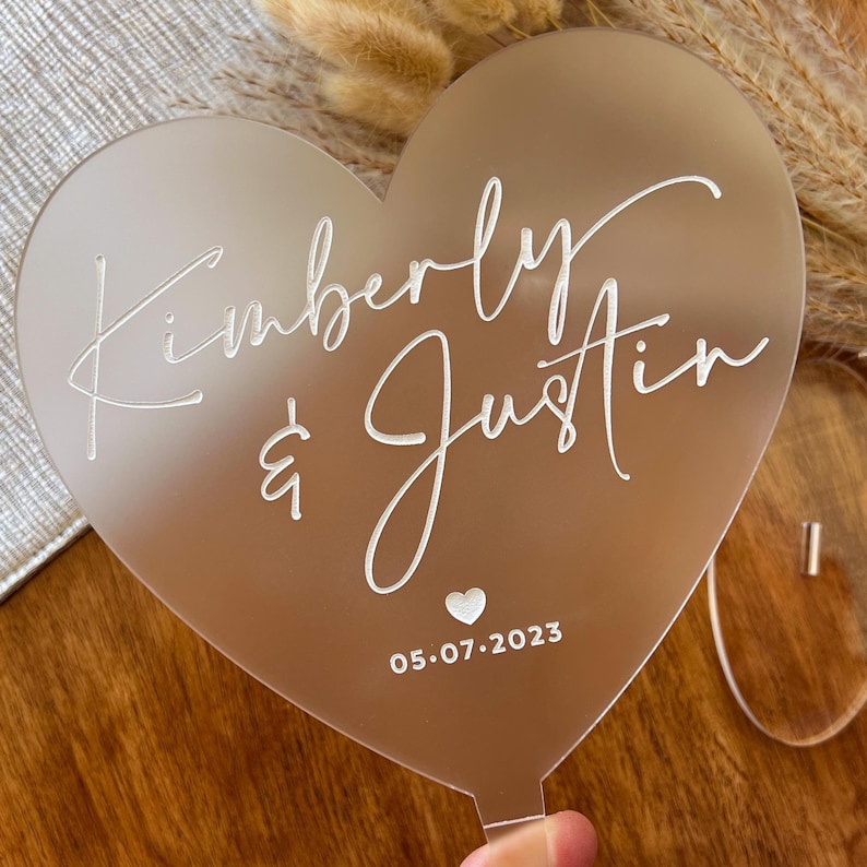 May include: A heart-shaped wedding cake topper with the names "Kimberly & Justin" and the date "05.07.2023" in white script. The topper is made of a translucent material.