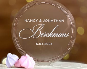 May include: A circular, reflective rose gold acrylic cake topper featuring the white printed text NANCY & JONATHAN, the script name Berckmans, and the date 6.04.2024. The topper has a smooth, glossy finish and a circular border design.