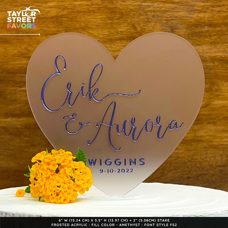 May include: A frosted acrylic heart-shaped wedding cake topper with the names "Erik & Aurora" in blue script. The names are above "Wiggins" and the date "9-10-2022". A cluster of yellow flowers sits in front of the topper.